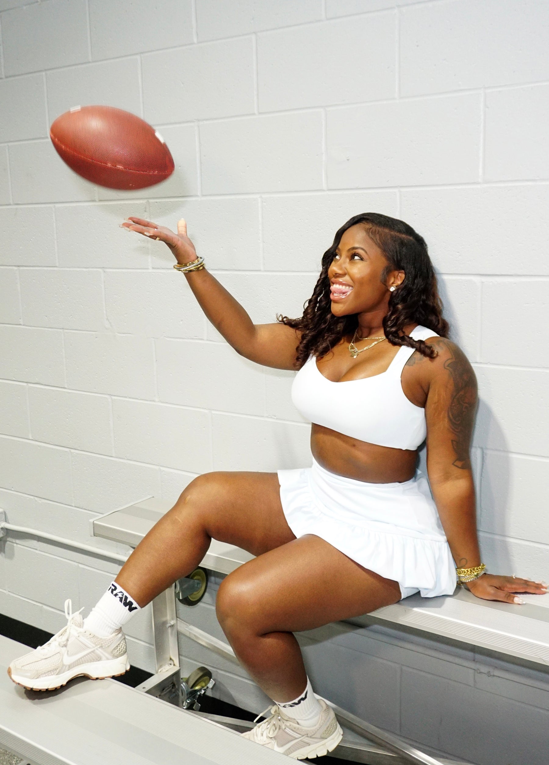 Woman in a white outfit with a football, sitting on a bench against a tiled wall.